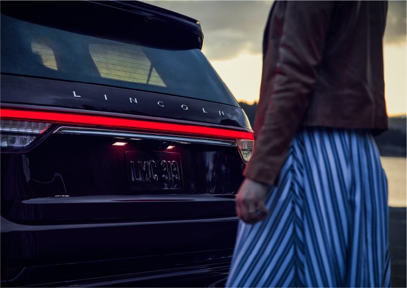A person is shown near the rear of a 2023 Lincoln Aviator® SUV as the Lincoln Embrace illuminates the rear lights | North Park Lincoln in San Antonio TX